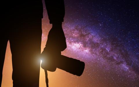 An up-close silhouette of a man holding a camera down by his side and is facing towards a purple and orange galaxy in the background.